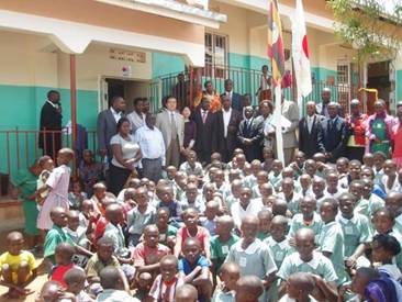 The ambassador standing with the district officials, school management, parents and the children in front of the classroom block
